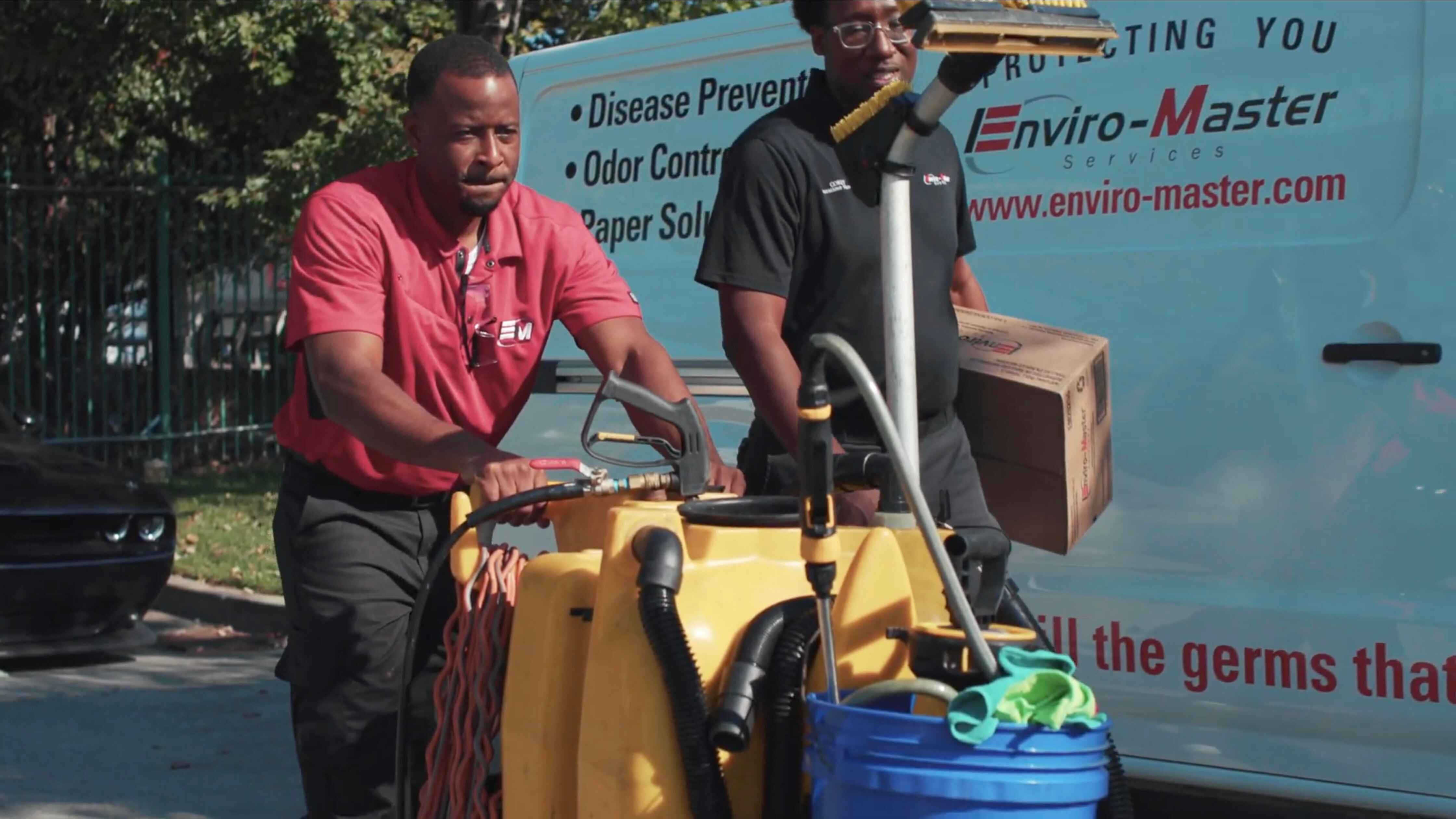 Two Enviro-Master technicians move cleaning equipment in front of an Enviro-Master vehicle.
